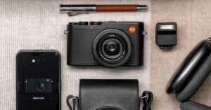 A flat lay photo features a Leica camera centered, surrounded by a pen, an external flash, a smartphone displaying a Leica camera image, a black leather camera case, and wireless headphones. The neutral background highlights the sleek, modern gear.