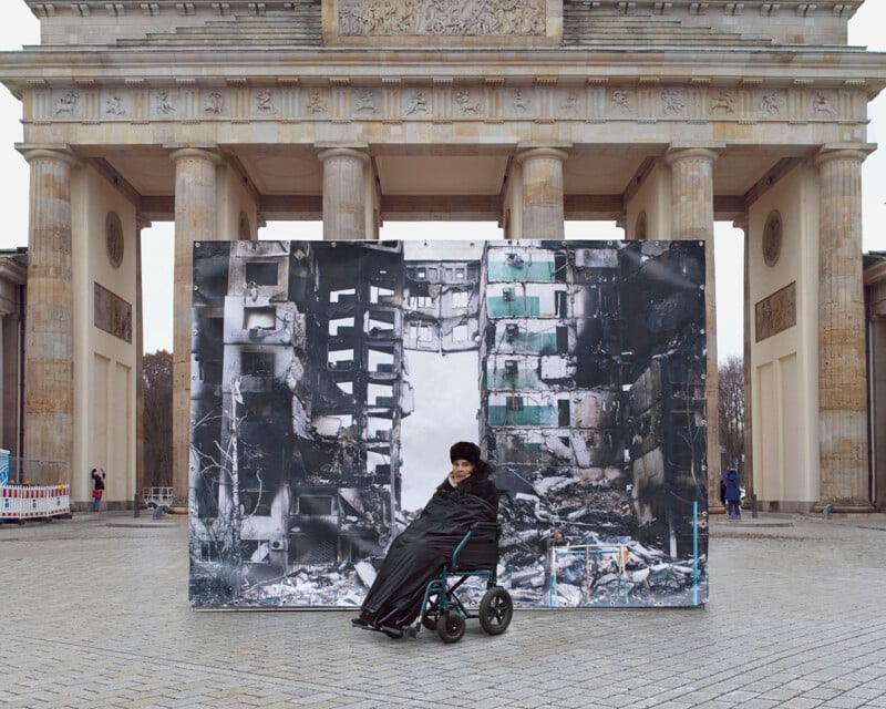 A person sits in a wheelchair, wearing a heavy coat and hat, positioned in front of a large black and white photograph of a bombed-out building. This scene is set in front of the Brandenburg Gate, a historic monument in Berlin.