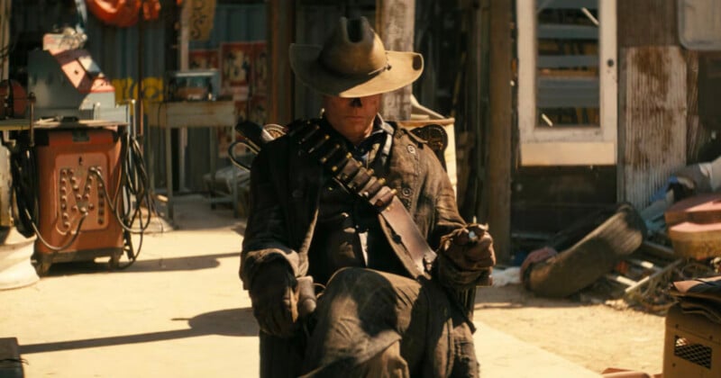 A lone man in a Western-style outfit sits cross-legged on a wooden chair in a dusty, old-fashioned town. He's wearing a wide-brimmed hat, long coat, and a bandolier full of bullets across his chest, exuding a rugged, cowboy demeanor.