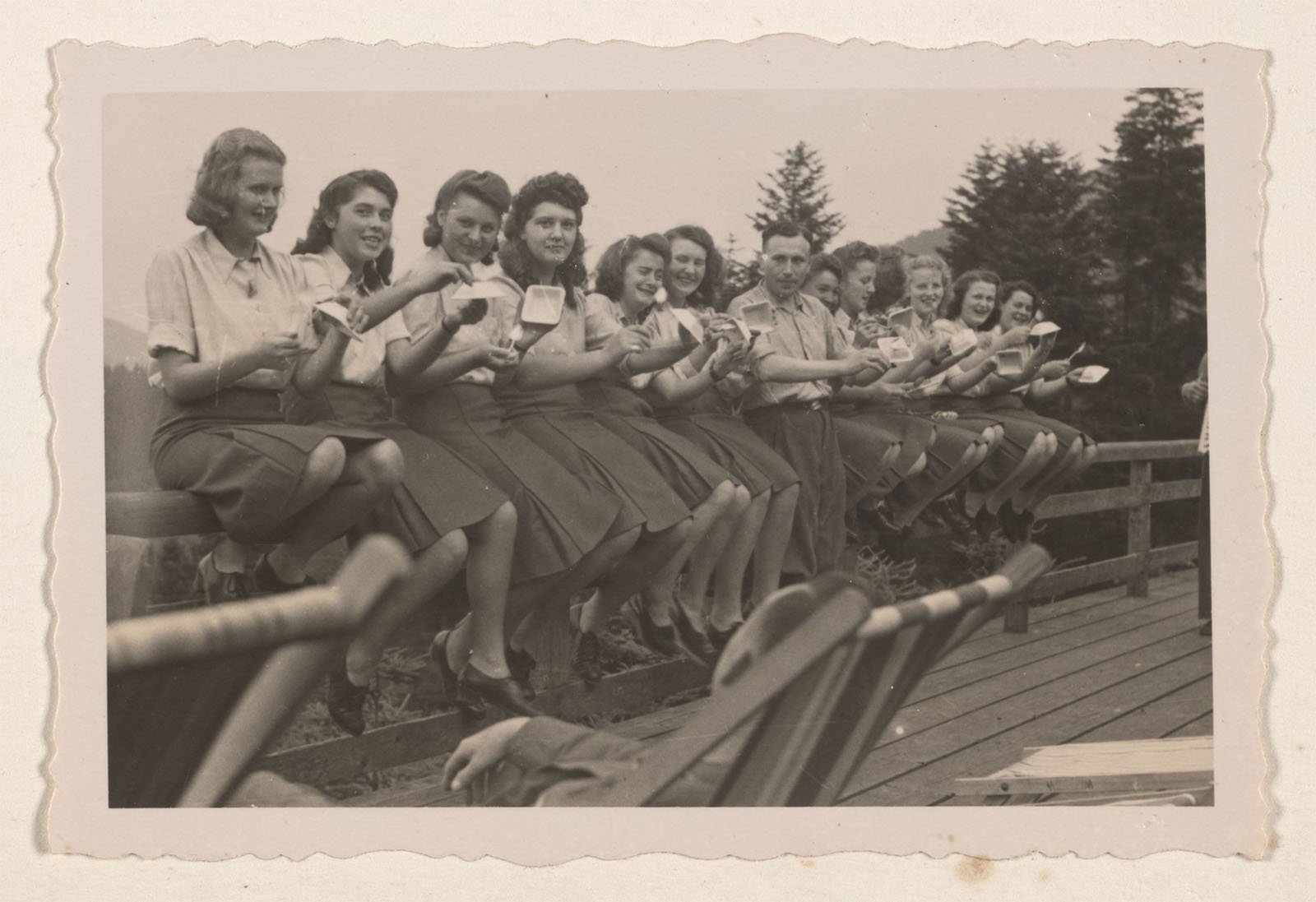 A vintage black-and-white photo shows a group of women and one man sitting in a row on a wooden fence, each holding a small cup and saucer. They are dressed in similar attire, likely from the mid-20th century. Trees and a wooden deck are in the background.