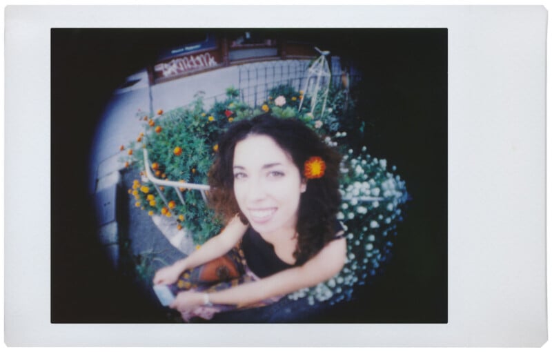 A smiling woman with curly hair stands outdoors in front of a garden of colorful flowers. The photo has a fisheye lens effect, causing a circular frame with a vignette around the edges. She is holding an object, possibly a book or a phone.