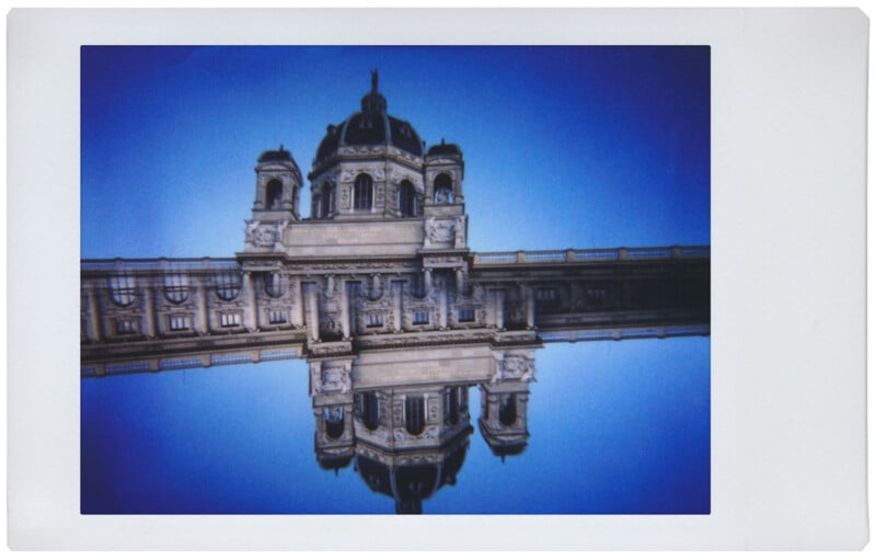 A Polaroid photo of a grand building with a central dome and two towers is reflected symmetrically in a clear blue body of water. The structure is set against a deep blue sky, creating a mirrored effect that blends architecture with its reflection.