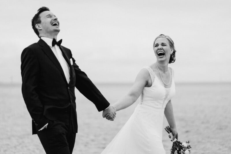 A joyful bride and groom laugh heartily while holding hands on a beach, wearing a wedding dress and a tuxedo, with a bouquet in hand and a vast ocean in the background.