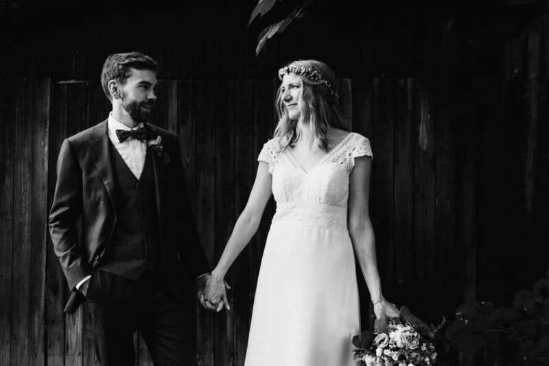 A monochrome image of a bride and groom holding hands. the bride, in a lace dress and floral headband, smiles at the groom who is dressed in a formal suit. they stand in front of a dark wooden backdrop.
