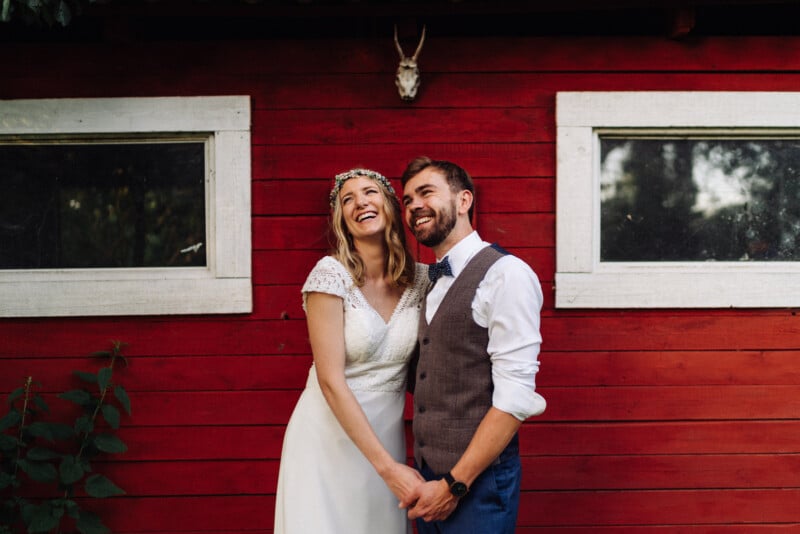 A joyful bride and groom laugh together in front of a red wooden cabin, the bride in a lace dress and floral crown and the groom in a vest and bow tie.