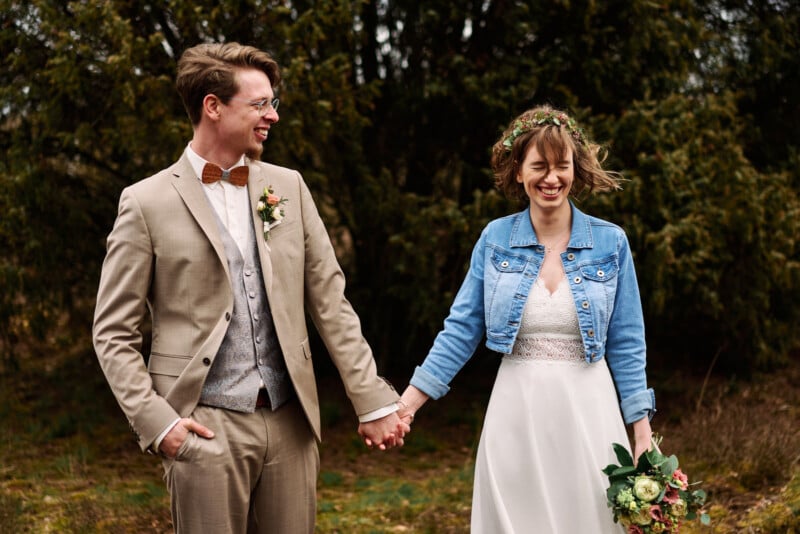 A joyful newlywed couple holding hands in a forest setting. the groom wears a tan suit with a bow tie, and the bride wears a white dress with a denim jacket and carries a bouquet.