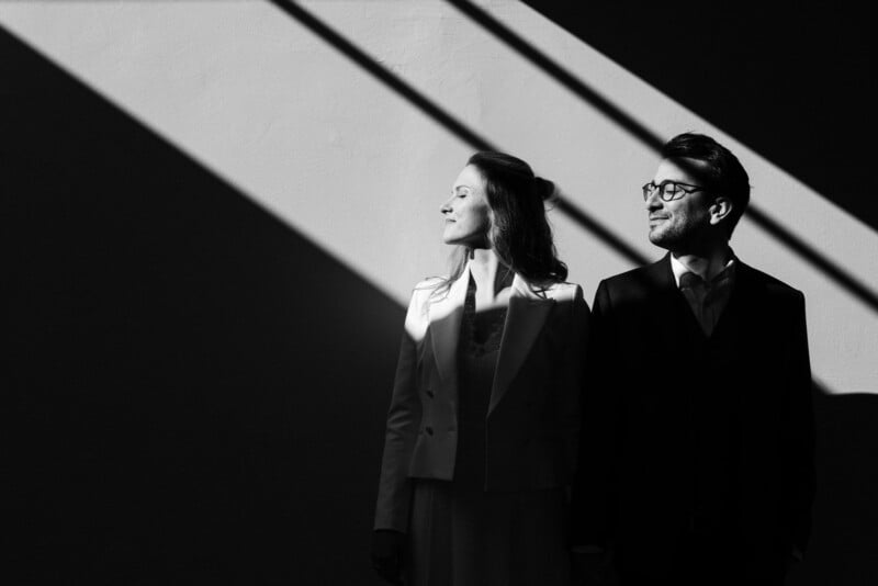A man and a woman dressed in formal attire, standing side by side, face profiles illuminated by diagonal light and shadows on a solid background.