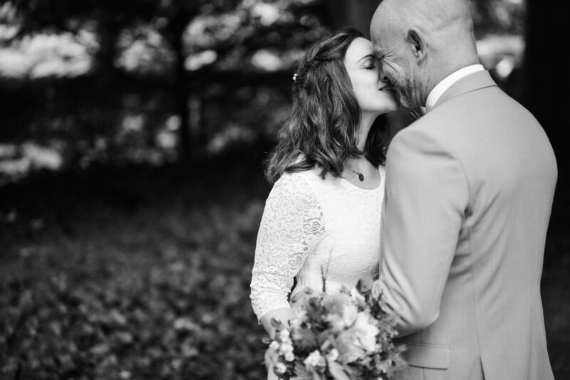 A black and white photo of a bride and groom intimately touching foreheads, smiling gently, amid a backdrop of soft-focus trees. the bride holds a bouquet and wears a lace dress.