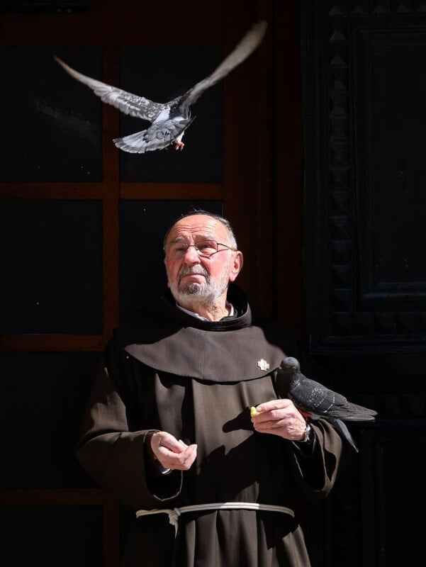 A man dressed as a monk or friar with a white beard and glasses stands outside, holding a pigeon in one hand and feeding another pigeon that is flying nearby. He is wearing a brown robe and the background is dark with part of a door visible.