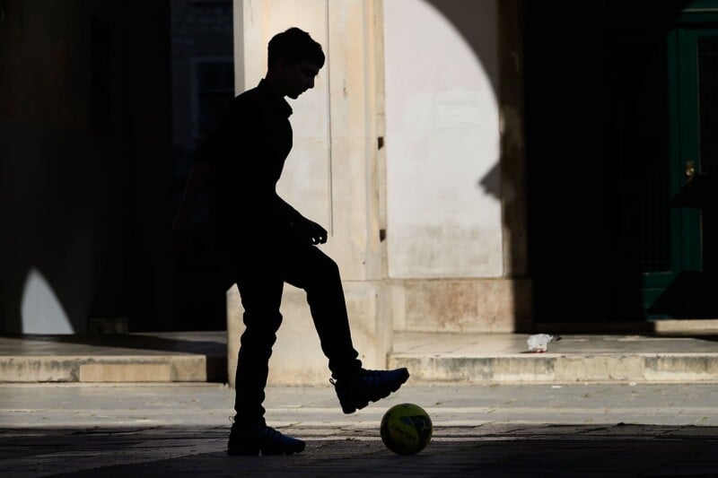 Silhouette of a person in a dark alleyway balancing a yellow soccer ball with one foot. The background features stone architecture with shadows and a partially visible green door. The scene is sunlit, creating contrasts between light and shadow.