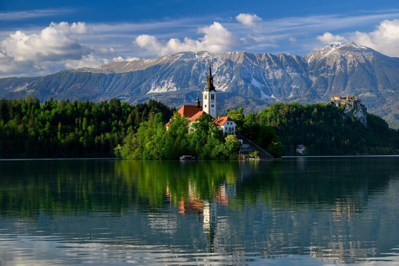 A picturesque island with a church sits in the middle of a tranquil lake, surrounded by lush greenery. Snow-capped mountains and a partly cloudy sky form a stunning backdrop, while the reflection of the scene shimmers on the water's surface.