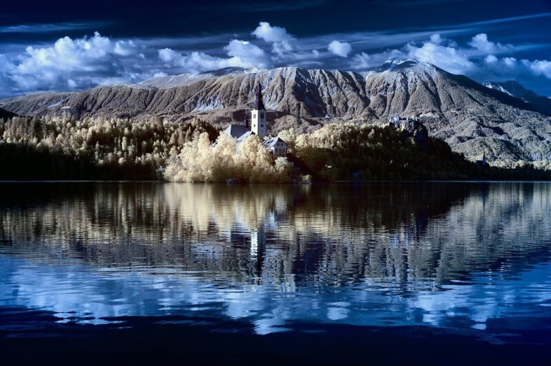 A serene lake reflects a picturesque island with a church, set against a backdrop of towering, snow-capped mountains under a partially cloudy sky. The scene is bathed in soft, ethereal light, creating a dreamlike atmosphere.