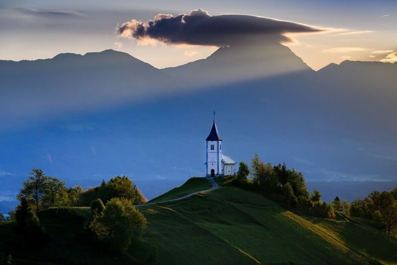 A white church with a tall steeple sits atop a lush green hill, with a winding path leading to it. Behind the church, a mountain range is bathed in golden sunlight as a unique cloud formation hovers above the peaks.