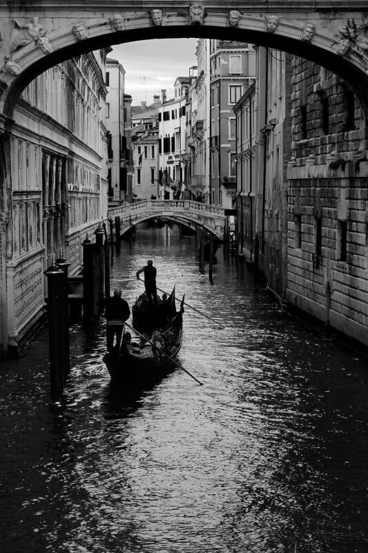 Black and white photo of a Venetian canal with a gondola piloted by a gondolier moving under a stone bridge. Historic buildings with intricate carvings flank the canal, creating a picturesque and classic Venice scene.