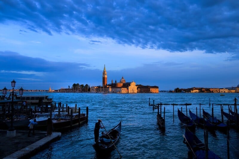 A view of a Venetian canal at dusk, showcasing gondolas moored at a dock with a backdrop of historic buildings, including a prominent church tower. The sky is filled with dramatic clouds, casting a serene blue and golden hue over the water and structures.