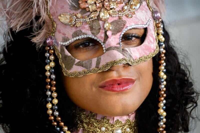 A person is wearing an ornate pink masquerade mask adorned with jewels, crystals, and pearls. The mask includes a feather on top and intricate gold detailing around the edges. The person also wears matching jewelry and has a slight smile.