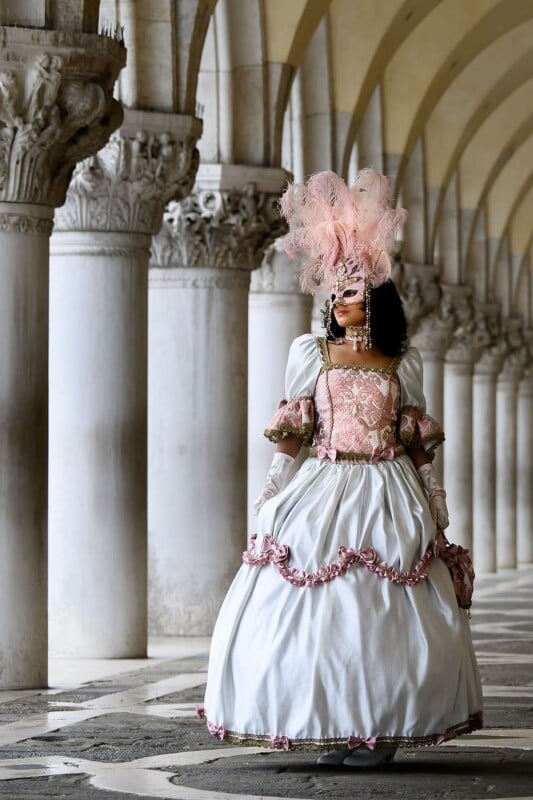 A person in an elaborate historical costume walks through a corridor of arched columns. The outfit features a large white and pink gown with gold accents, a pink lace corset, and a feathered mask and headdress, evoking a Venetian carnival or royal ball.