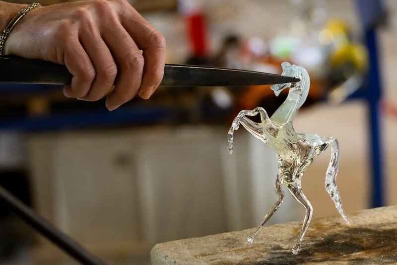 A close-up of a hand using tongs to shape a delicate, clear glass sculpture of a rearing horse. The sculpture sits on a work surface, and the background shows a slightly blurred workshop environment.
