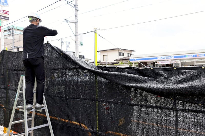 A worker wearing a helmet stands on a ladder, adjusting a black mesh barrier near an outdoor construction site. Buildings and a Lawson convenience store are visible in the background, with electrical wires crossing overhead on a cloudy day.