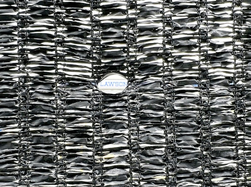 A textured, woven metallic surface with a shiny, reflective appearance. There is a small, round, white label with partially visible blue text “AWSON” that is partly obscured by the metallic material. The weave is tightly knit in a grid pattern.