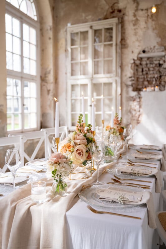 A beautifully set dining table with elegant white tablecloths and floral centerpieces featuring roses and greenery. Tall white candles in gold holders, clear glassware, and white plates with gold cutlery add a sophisticated touch. The background features large windows and a rustic wall.