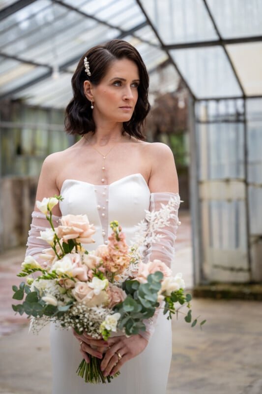 A bride in a strapless white wedding gown with sheer off-the-shoulder sleeves holds a bouquet of white, peach, and greenery flowers. She stands in a rustic greenhouse, looking thoughtfully to the side. Her dark hair is styled in loose waves, adorned with a hairpin.