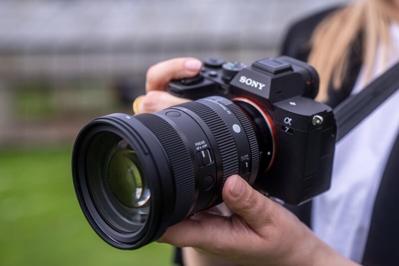 A close-up of a person holding a Sony digital camera with a large lens attached. The camera is black, and the person is gripping it with both hands, standing outdoors with a blurred background.