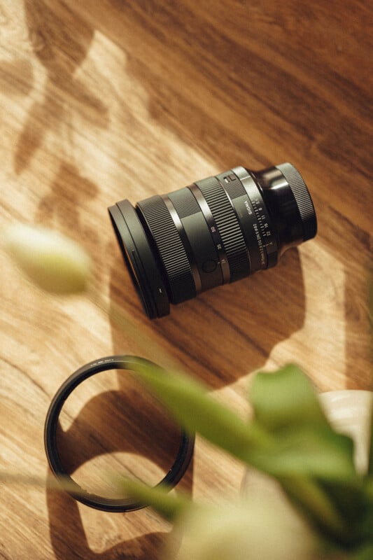 A camera lens rests on a wooden surface with soft light and shadows. A detached lens cap lies nearby, and an out-of-focus plant is in the foreground, creating a serene atmosphere.