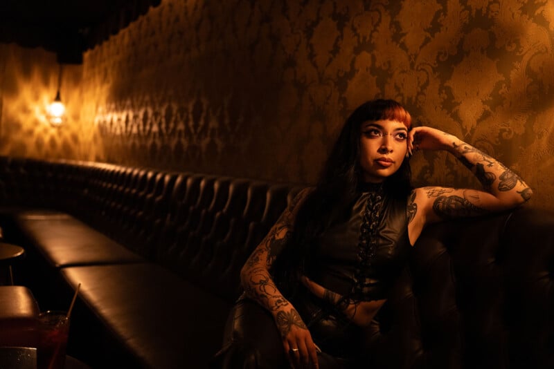 A woman with long black hair and tattoos sits on a black leather couch in a dimly lit room with ornate wallpaper. She wears a black sleeveless top and rests her arm on the back of the couch, looking thoughtfully into the distance.