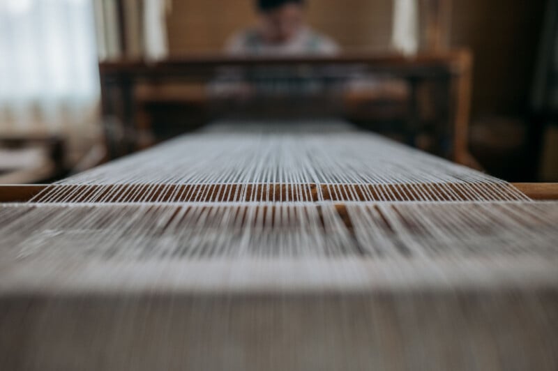 Close-up view of threads on a traditional weaving loom with a blurry figure of a person operating the loom in the background. The image focuses on the aligned threads stretching towards the weaver, capturing the intricate details of the weaving process.