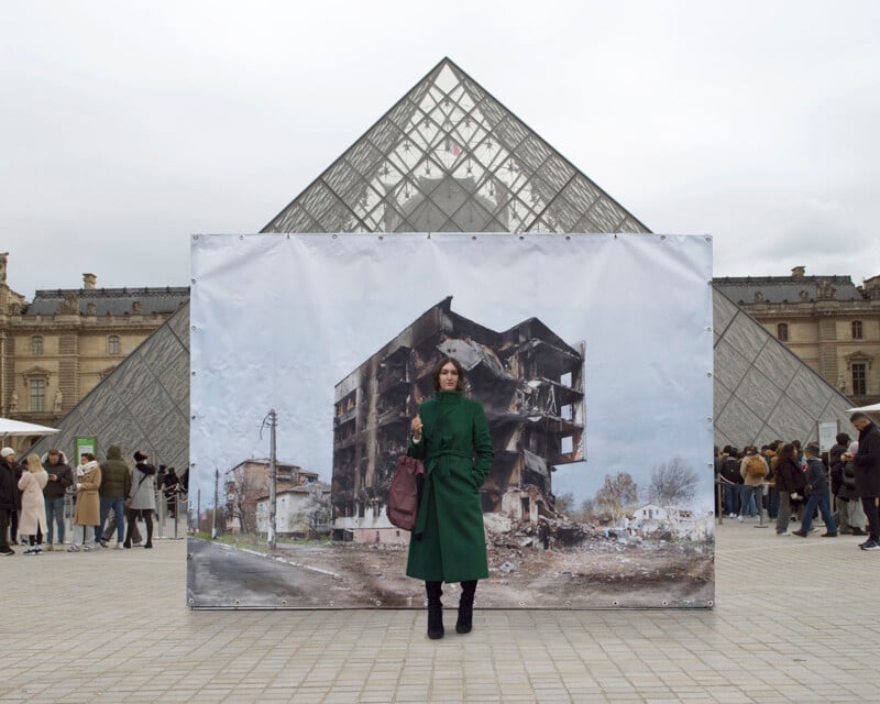 A person in a long green coat stands in front of an outdoor display depicting a damaged building. The display is set in front of the Louvre Pyramid, with several people and part of the museum's architecture visible in the background.