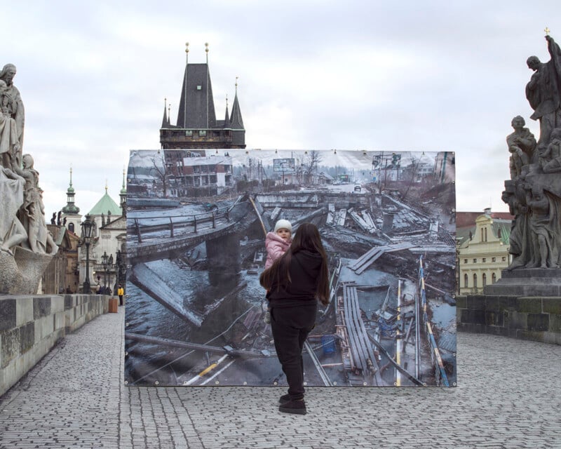 A person in black clothing carries a child while standing before a large, outdoor photograph of a destroyed bridge. The scene is set on a historic stone bridge lined with statues. Tall, pointed towers rise in the background under an overcast sky.