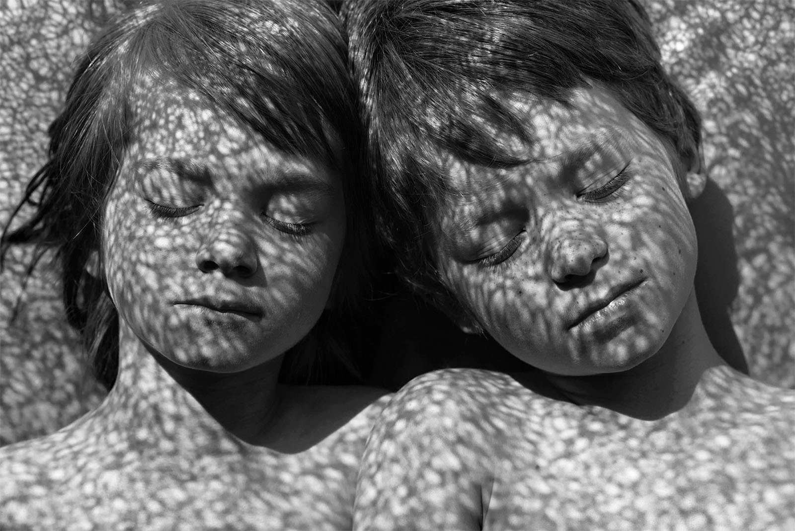 Black and white photograph of two children lying side by side with closed eyes. Dappled light filters through a textured surface above them, casting intricate shadows on their faces and bodies. Their expressions are peaceful and relaxed.