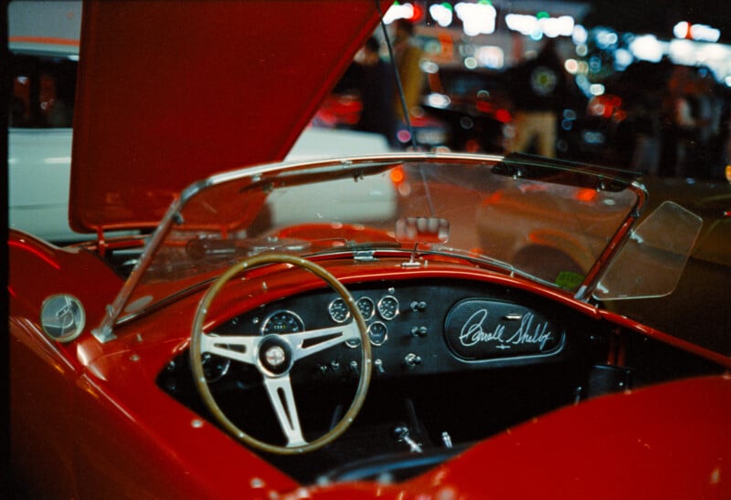 Close-up of the interior of a vintage red sports car with the hood raised. The steering wheel is prominently displayed along with a dashboard that features multiple gauges and an autograph reading "Carroll Shelby." The background is blurred with other cars and people.