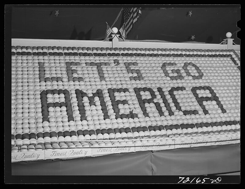 A display made up of numerous eggs arranged to spell "LET'S GO AMERICA." The display is part of a larger booth, decorated with stars and stripes at the top and a patterned banner at the bottom that reads "Finest Quality." An American flag is visible in the background.