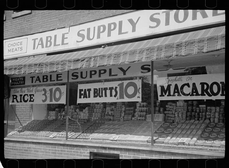 Black-and-white photo of a storefront window display for "Table Supply Store." The display includes signs for "Rice 3 10¢," "Fat Butts 10¢," and "Spaghetti or Macaroni" alongside shelves stocked with various cans and packages.