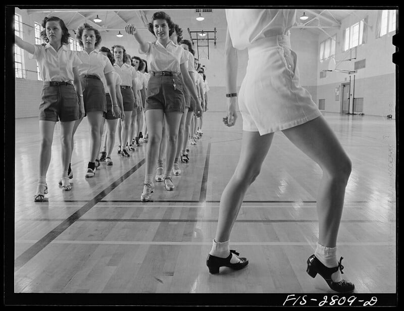 A black-and-white photo from the 1940s showing a line of young women in a gymnasium practicing a dance routine. They are dressed in shorts and blouses, with their arms extended forward and marching in sync. The gym has high ceilings and tall windows.