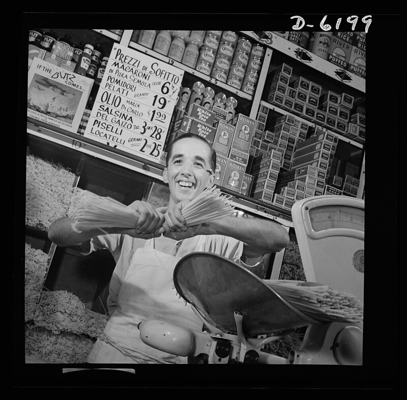 Black and white photo of a man smiling, holding bundles of spaghetti in a grocery store. Shelves behind him are stocked with various Italian food products, including pasta and sauces. A scale is visible on the counter in front of him.
