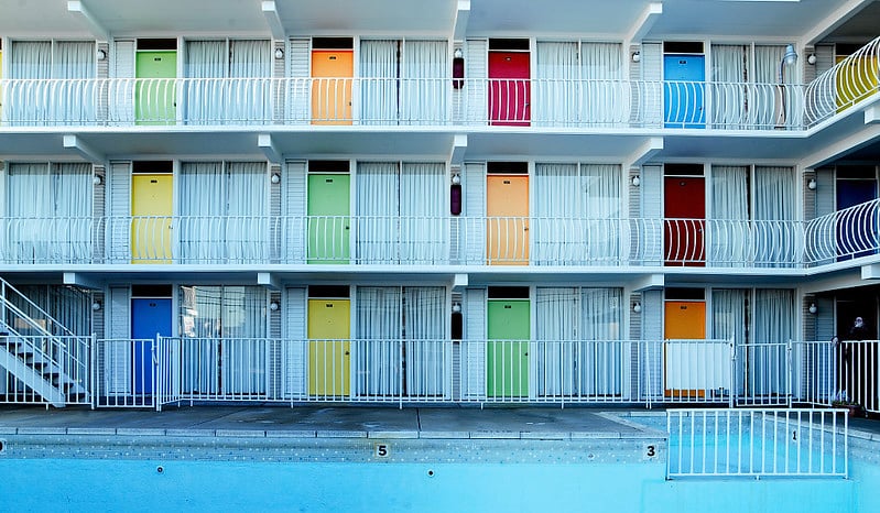 A three-story building with colorful doors in shades of yellow, green, blue, and red arranged in a grid pattern. The building features white balconies with railings and a swimming pool in the foreground. Stairs are visible on the left side leading to the upper floors.