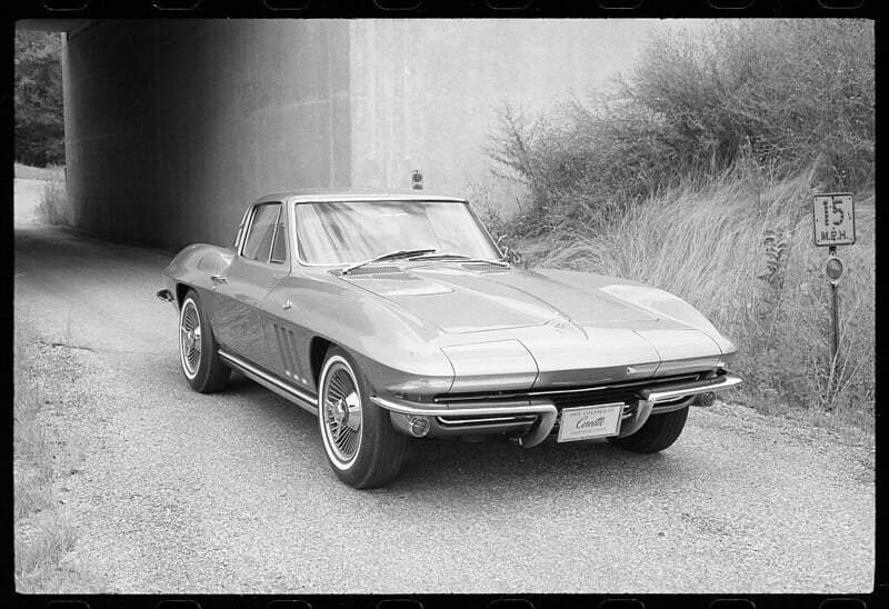 A black-and-white photo of a classic Chevrolet Corvette parked under a bridge. The car has a sleek, vintage design with prominent front grilles and detailed rims. A sign indicating a speed limit of 15 mph is visible on the right side of the road.
