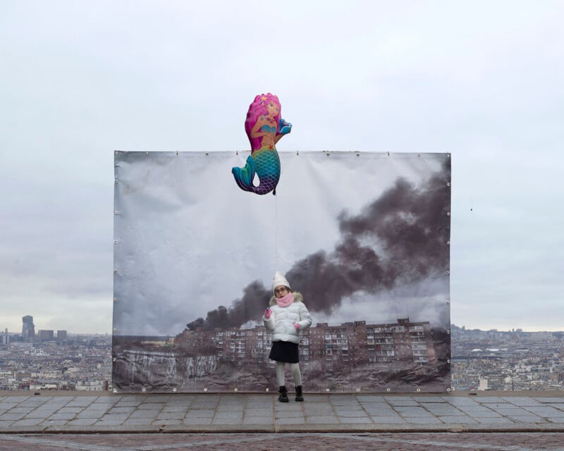 A young child in a white winter coat and hat stands in front of a large poster depicting a city with buildings and black smoke. The child holds a colorful seahorse-shaped balloon against a cloudy sky backdrop.