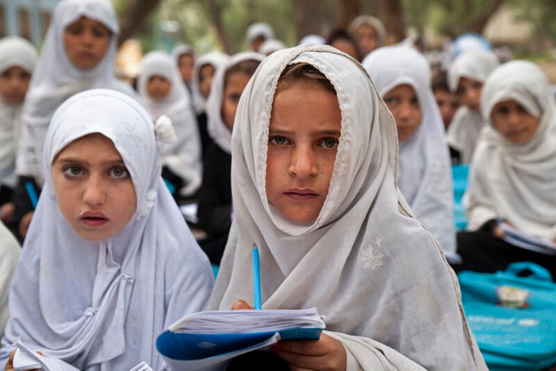 A group of young girls wearing white headscarves and black outfits sit outdoors on the ground. They hold notebooks and pencils, appearing attentive and focused. The background shows blurred trees and more children.