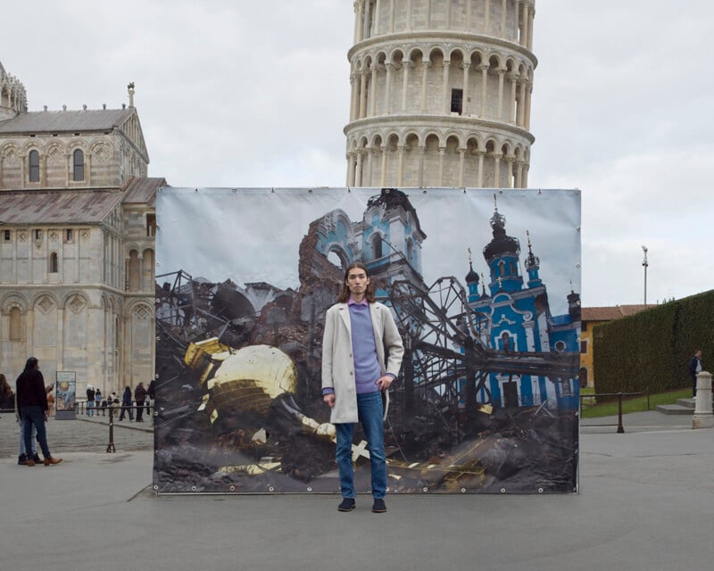 A person standing in front of a large banner displaying a photograph of a destroyed building set against the backdrop of Pisa's Leaning Tower and the Cathedral. The person is wearing a coat and jeans, posing with one hand on their hip. The sky is overcast.