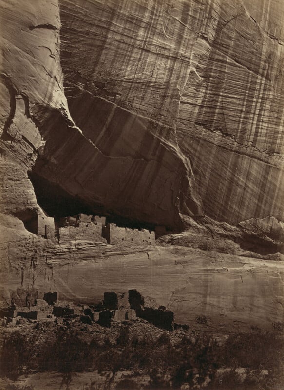 An ancient cliff dwelling is nestled into the side of a steep, massive rock face. There are multiple stone structures, some partially in shadow. The rock wall above the dwellings shows vertical striations, giving a textured appearance. Sparse vegetation is visible below.
