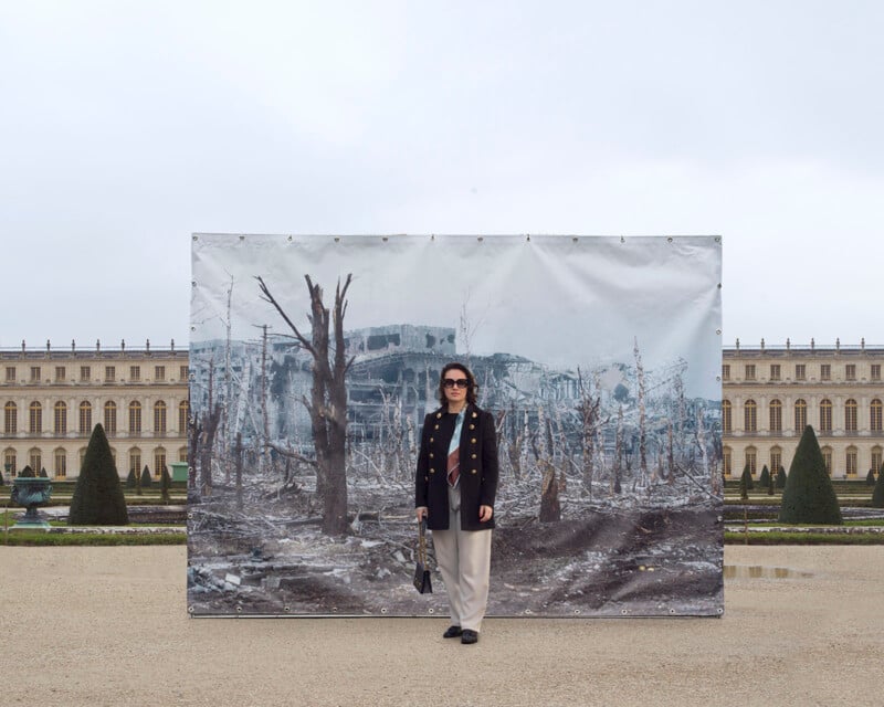 A person wearing a black coat and sunglasses stands in front of a large outdoor photograph. The photograph depicts a barren, war-torn landscape with destroyed buildings and leafless trees. Behind the person and photo, there is an ornate historical building and manicured gardens.