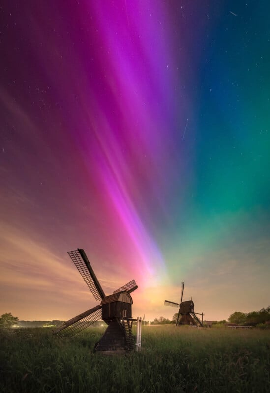 A vibrant aurora borealis illuminates the night sky with pink and green hues above two traditional windmills in a grassy field.