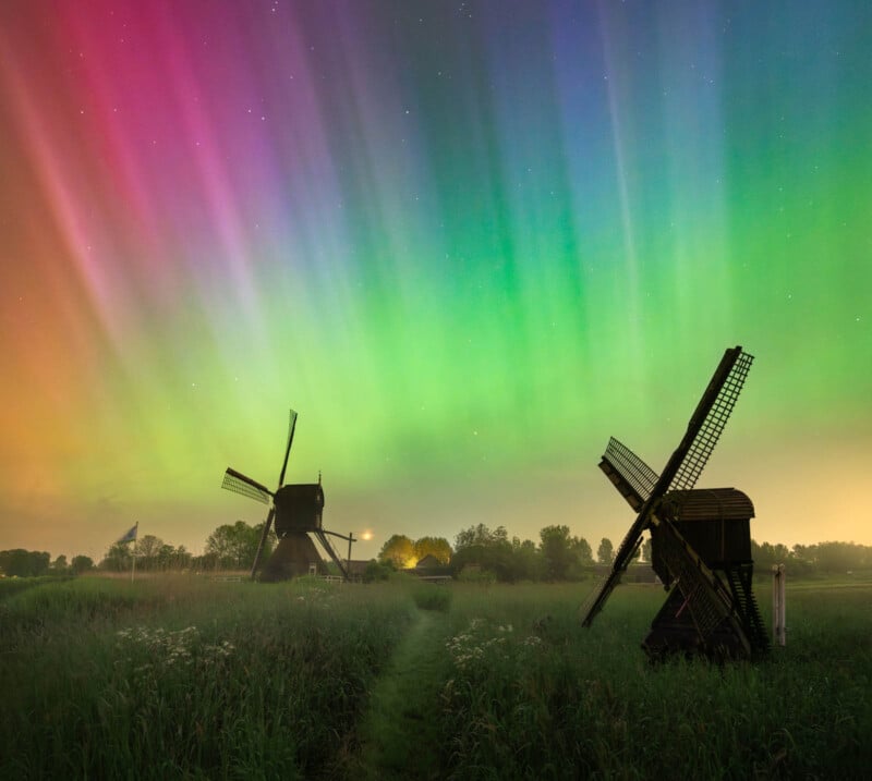 Vibrant aurora borealis above traditional Dutch windmills in a serene meadow at night, with a clear starry sky and colorful green and pink lights illuminating the scene.
