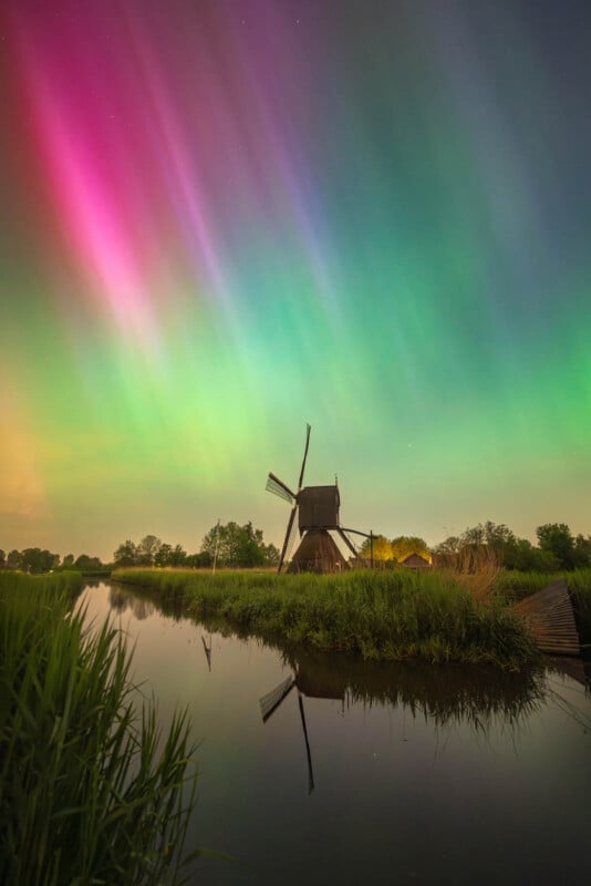 A vibrant display of the northern lights casting pink and green colors in the sky above a traditional windmill reflected in a calm river surrounded by lush greenery at night.