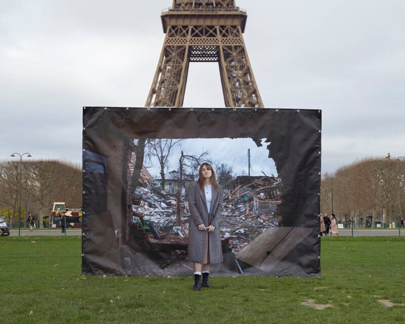 A woman stands in front of a large photo backdrop depicting the aftermath of destruction, with debris and ruins. The backdrop is set up in front of the Eiffel Tower in Paris, contrasting the serene park setting with the chaotic scene in the photo.