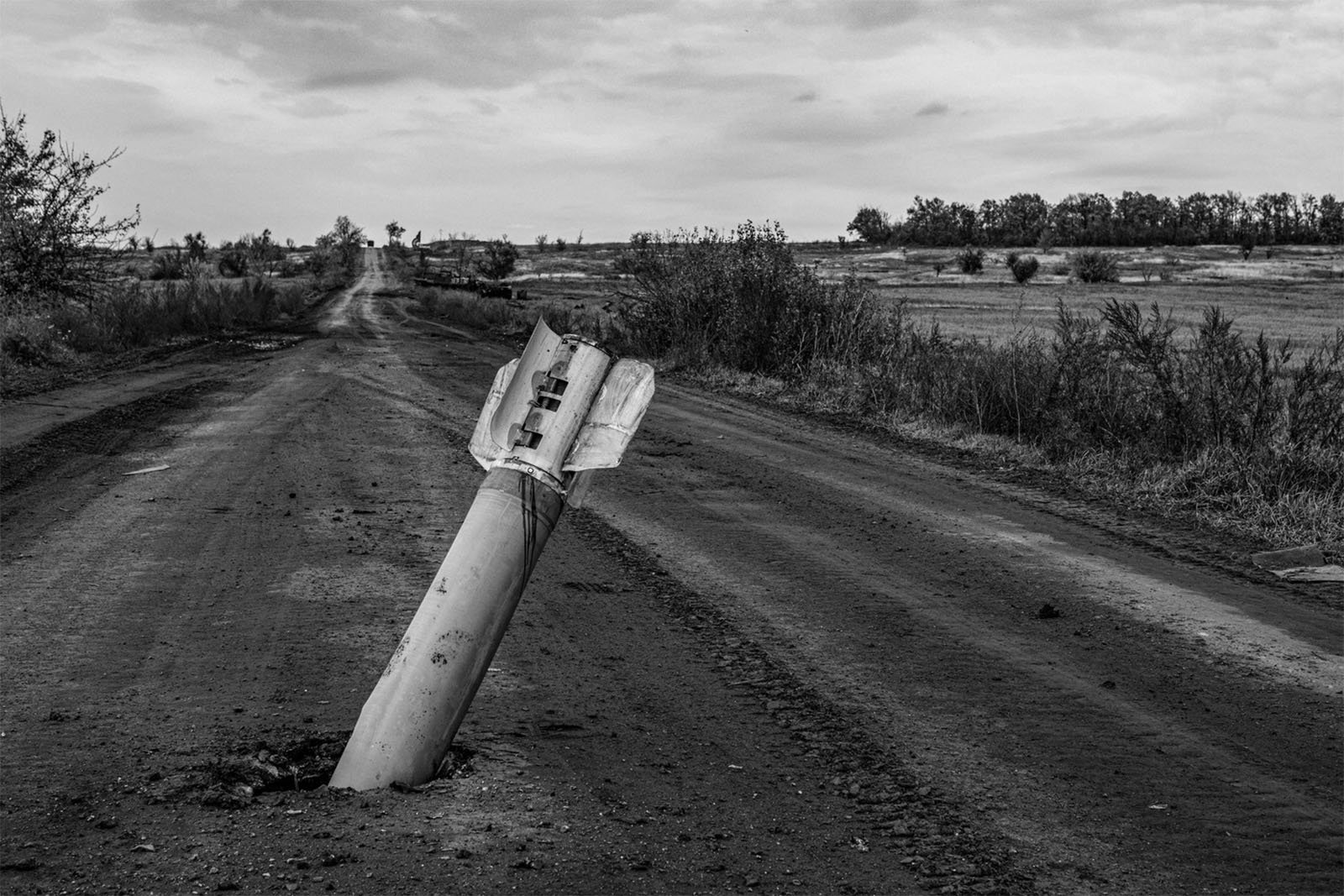 Black and white photo showing a large, unexploded missile lodged in the center of a remote dirt road. The landscape is barren with sparse vegetation and a cloudy sky. Trees and shrubs line the road's edges in the background.